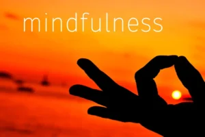 Mindfulness written in orange and yellow sunset sky with a person holding their pointer finger and thumb together as if they were meditating | What is Mindfulness | Breaking Dawn Therapy