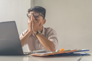 Man Sitting at a Table With a Computer and Folders With His Hands Covering His Face Pushing Up On His Glasses From Being Stressed | Overcoming Stress With Cognitive Behavioral Therapy | Breaking Dawn Therapy