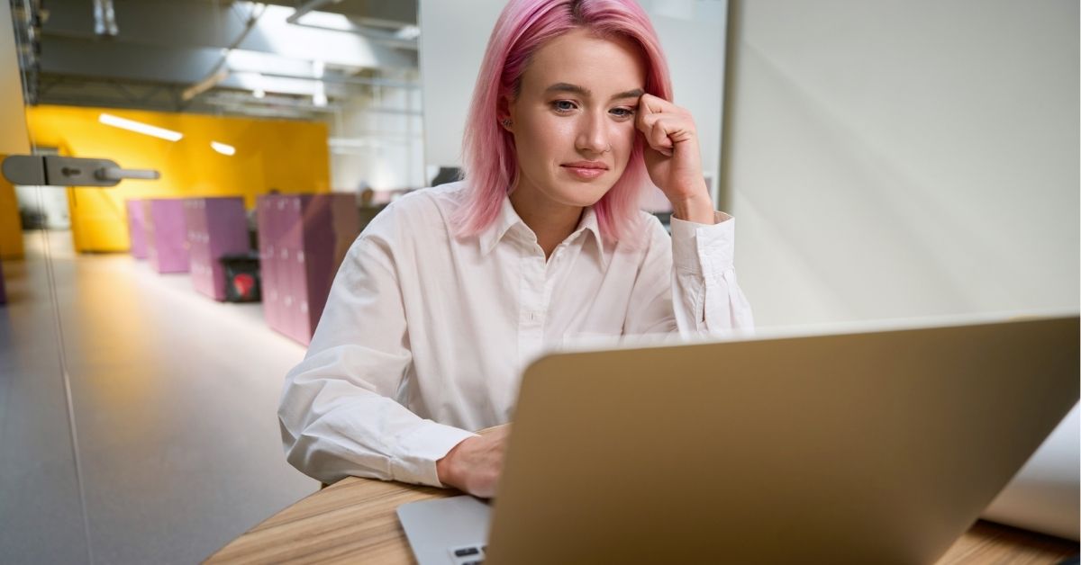 Woman with pink hair works thoughtfully on a laptop at a desk in a modern office environment.