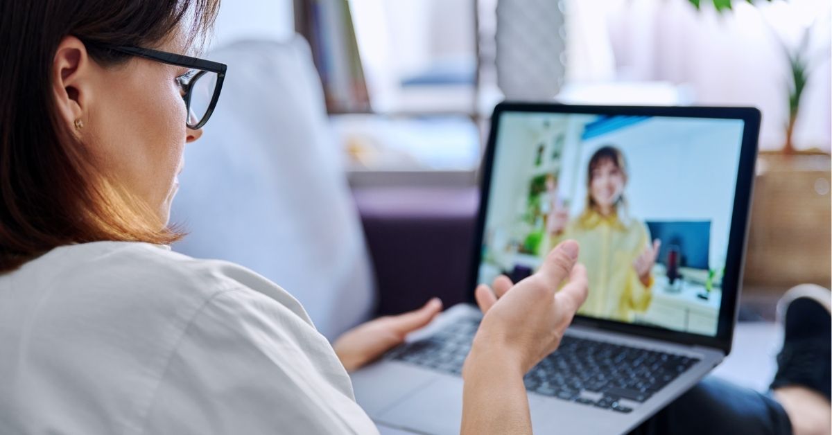 Woman wearing glasses gestures while speaking during a video call on a laptop in a relaxed home setting.