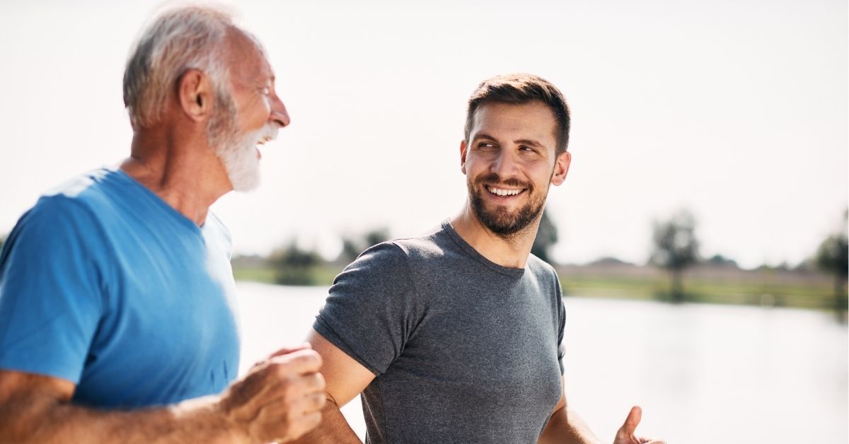 An older man and a younger man smile at each other while jogging together beside the water on a bright day.