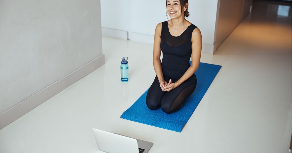 Smiling woman kneels on a blue yoga mat facing a laptop during an online yoga or fitness session at home.